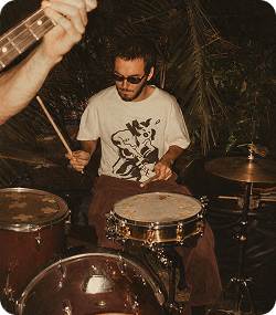 Músico con gafas de sol tocando la batería con energía en un espacio interior con plantas al fondo.